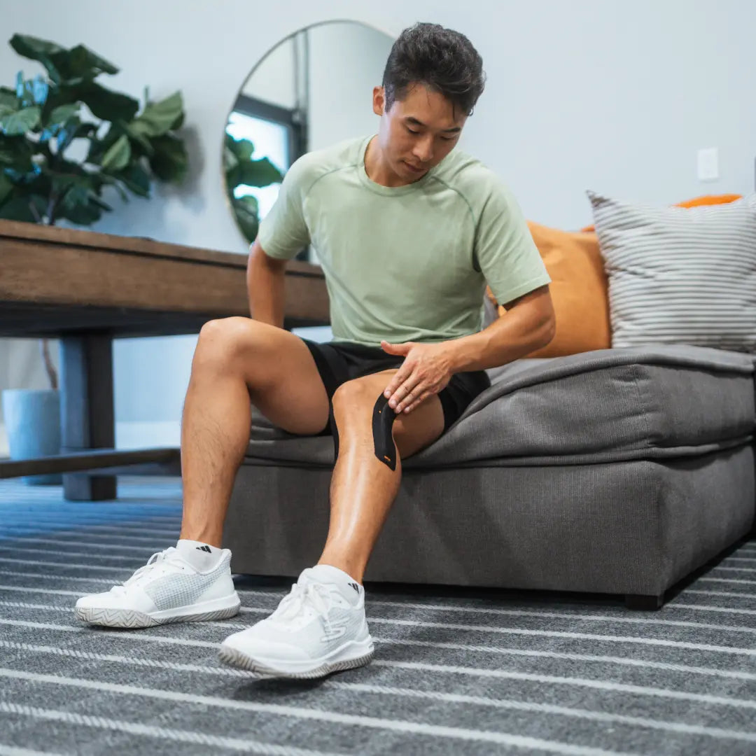 Person seated on a couch applying KT Tape to the lower leg, demonstrating self-application of kinesiology tape for muscle support and recovery at home.